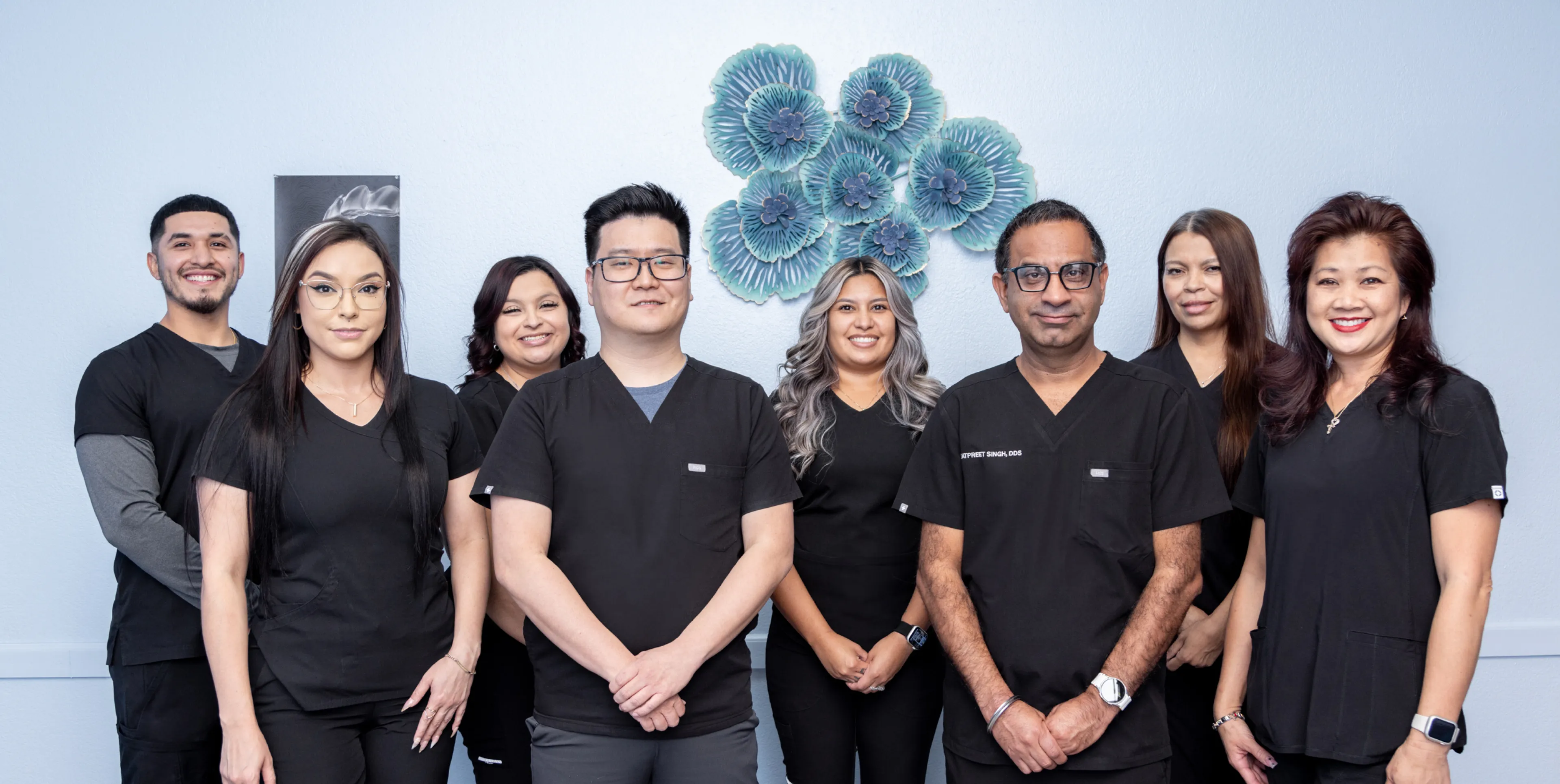 Group of eight medical professionals in black scrubs standing in a row and smiling against a light blue wall with decorative blue flowers.