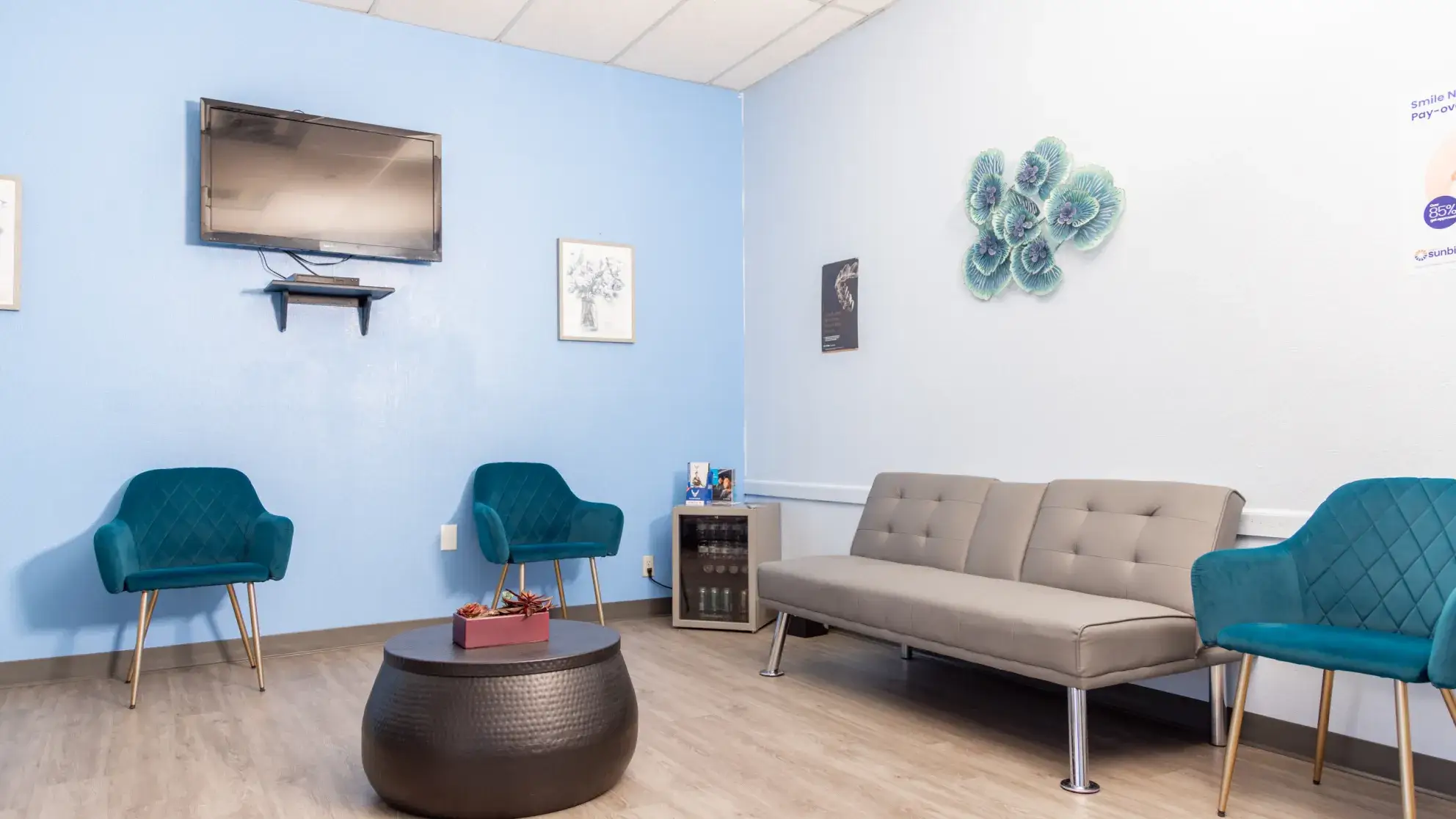 Modern waiting room with blue and gray walls, teal chairs, a gray sofa, a round black coffee table, and a wall-mounted TV.